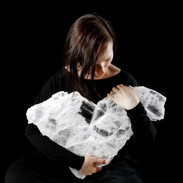 Laura Metz holding a large selenite crystal heart, eyes closed in reverence, black background